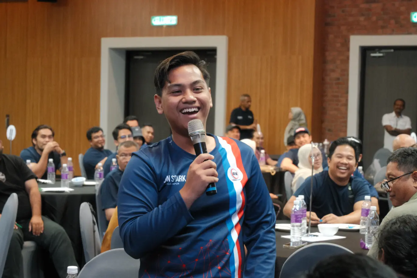 An early-career participant speaking into a microphone before a seated cohort during a Protege programme session